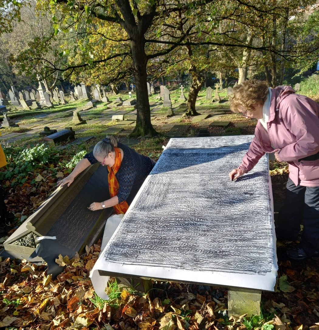 Two women creating gravestone rubbings at a table in St Mary&rsquo;s Churchyard, Prestwich, with rows of headstones behind them