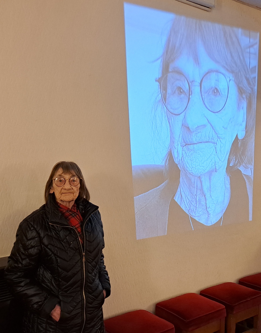 An elderly woman in a colourful scarf seated in front of canvases from the Portraits Exhibition, including a shopfront portrait and an elderly couple