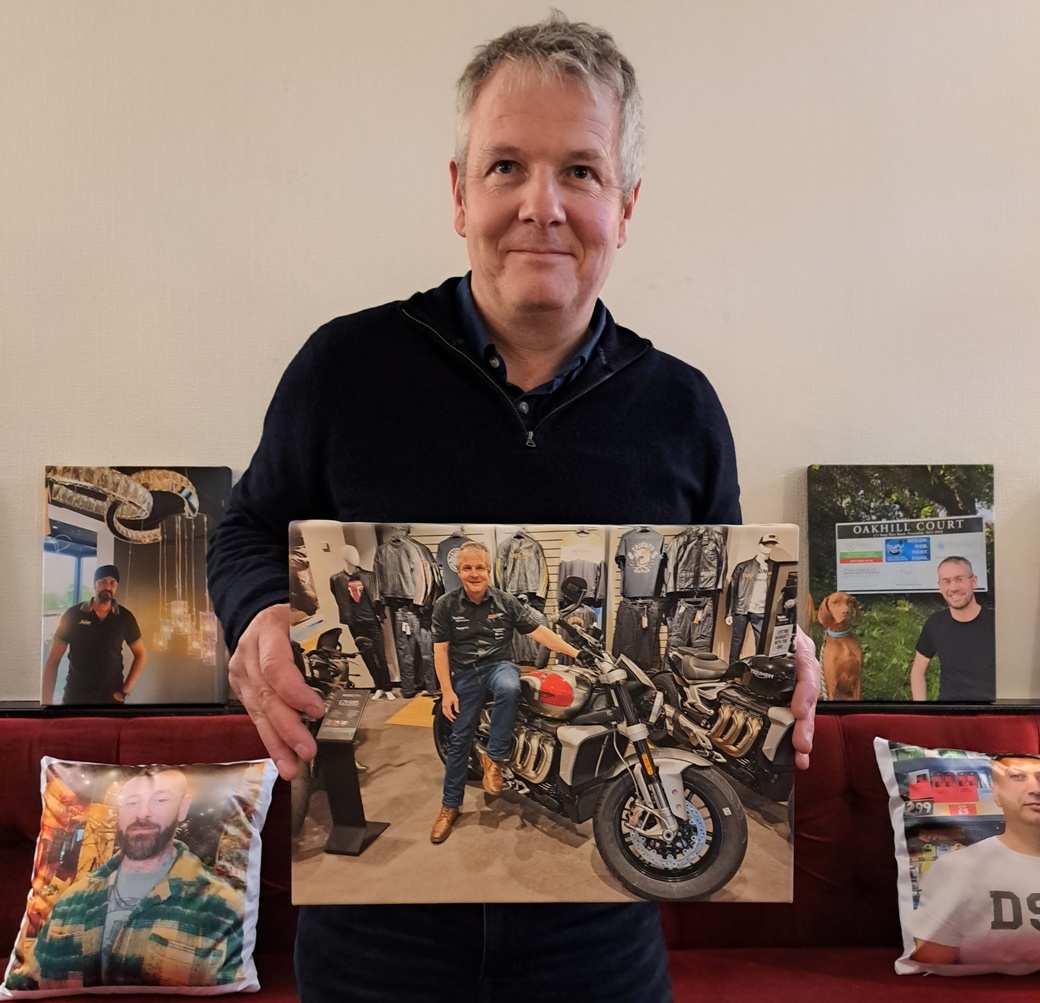 A man holding a canvas portrait of himself in a motorbike shop, with further canvases from the Portraits Exhibition and cushions behind him