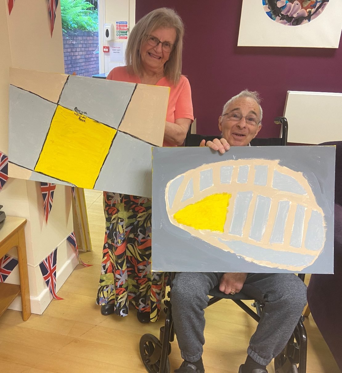 Two workshop participants holding up two canvases, a grey and yellow tiled pattern and a grey striped shield shape with a yellow triangle
