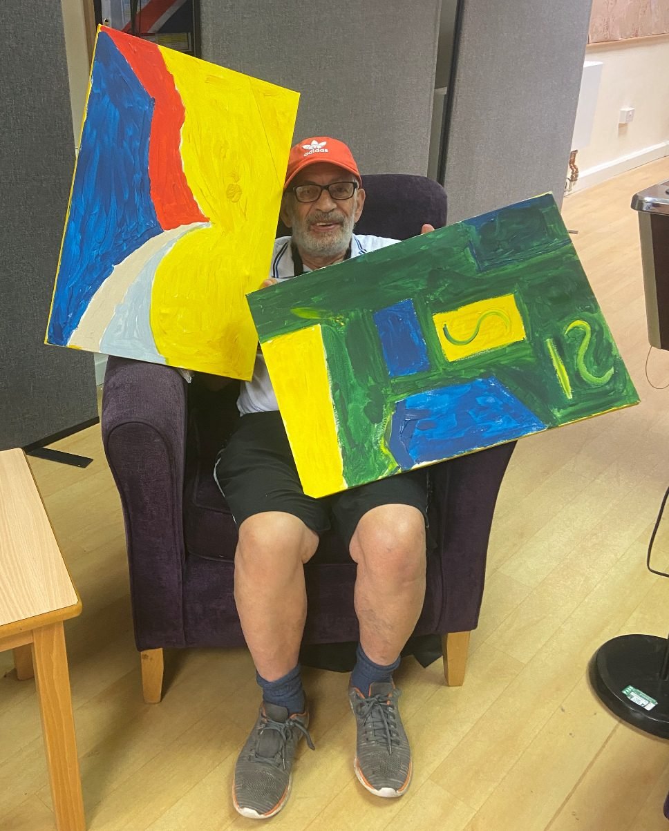 A workshop participant in a red cap holding up two abstract canvases in Prestwich Palette colours, one a blue, red and yellow flag-like design, the other a green and blue geometric pattern