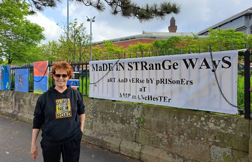 Louise Garman in a Bury New Road hoodie beside the &lsquo;Made in Strange Ways&rsquo; banner for the HMP Manchester prison art exhibition