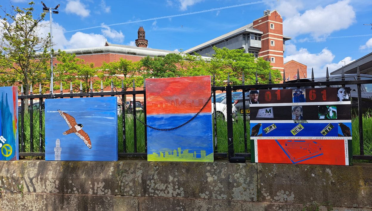 Canvases chained to the prison railings featuring a kestrel, a Manchester skyline in red and blue, and a collage board reading &lsquo;01 61&rsquo;