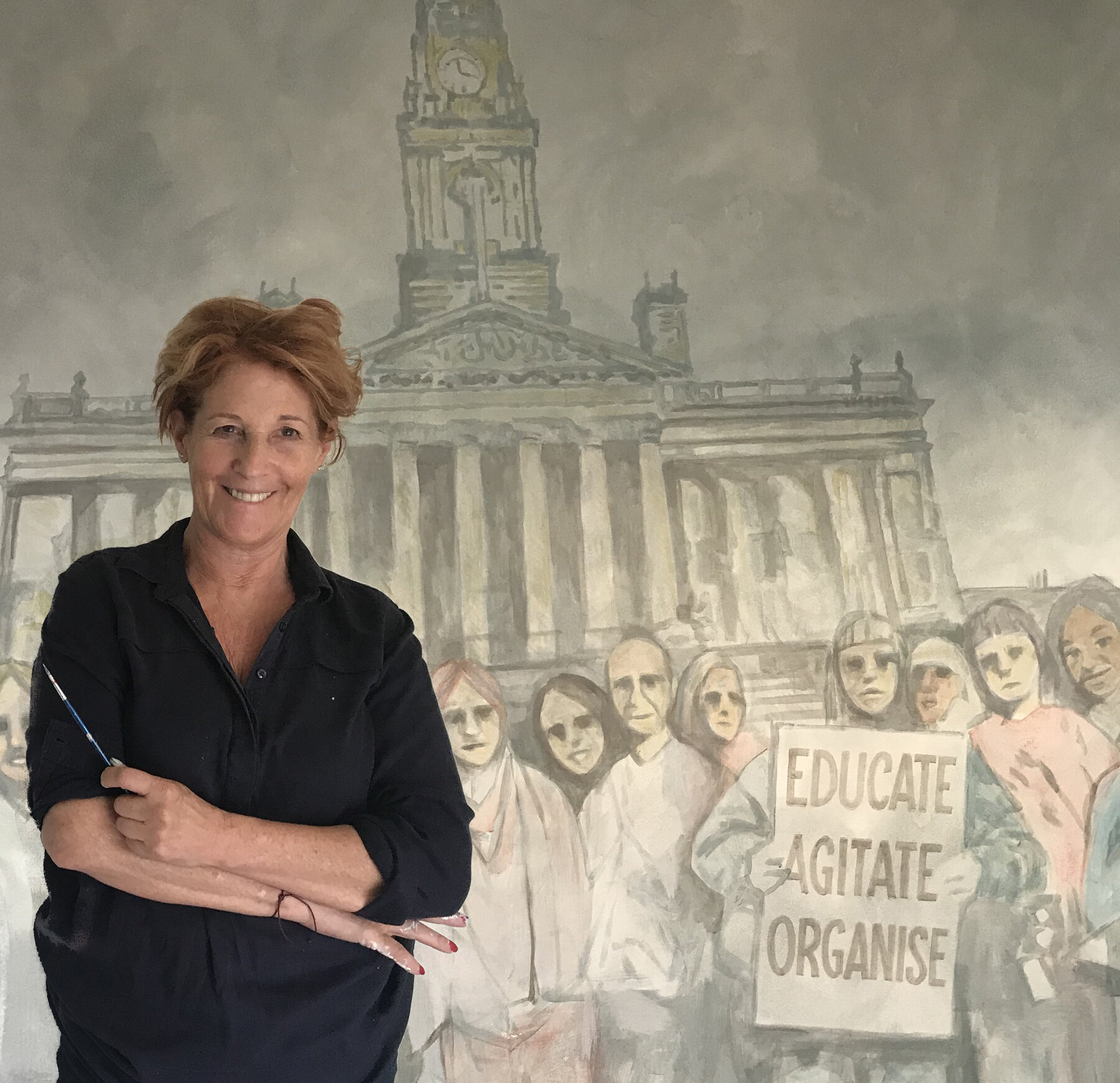 Louise Garman holding a paintbrush in front of her mural of a protest march past a town hall clock tower, with a placard reading Educate, Agitate, Organise