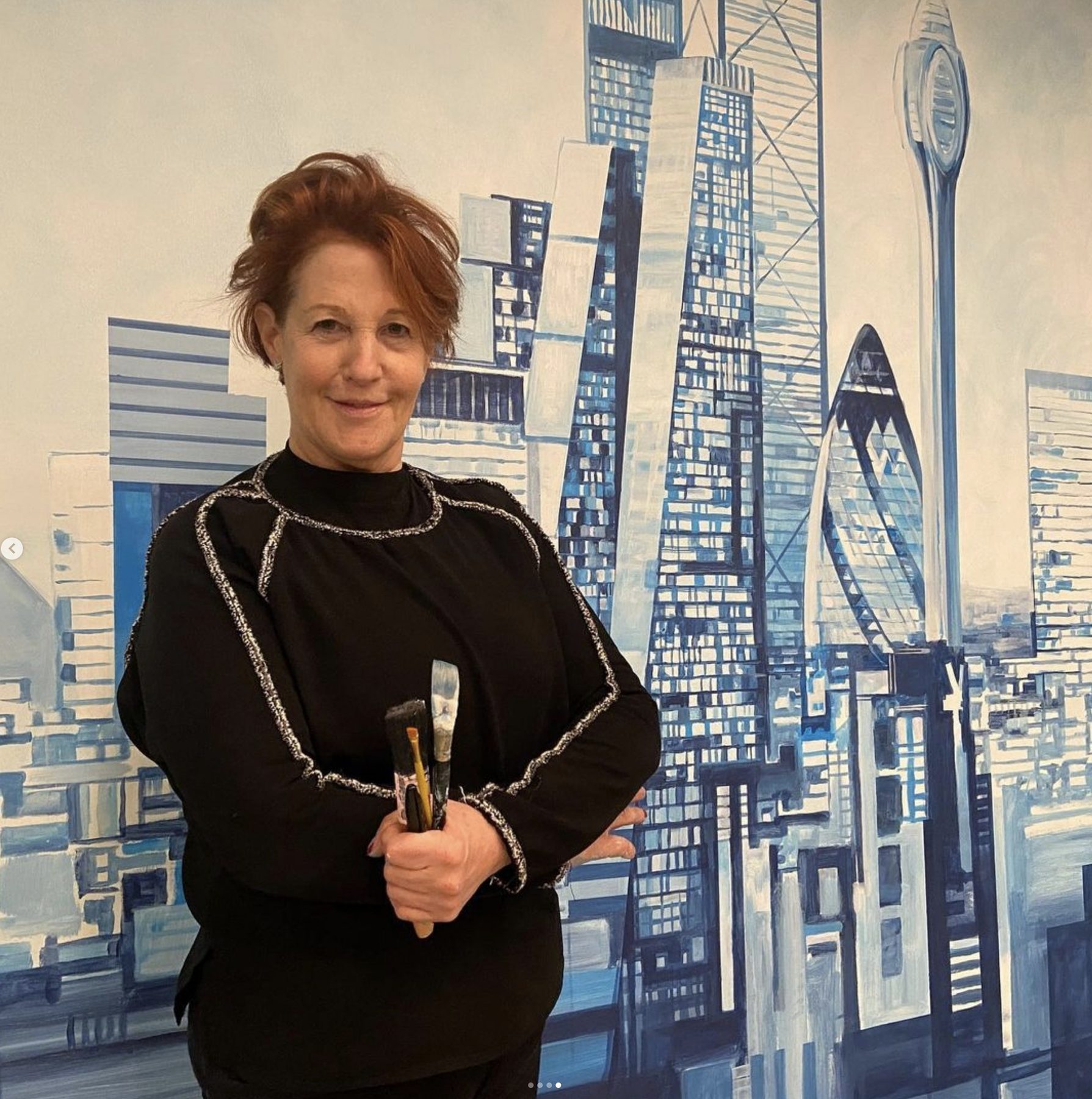 Louise Garman holding paintbrushes in front of the London skyline mural