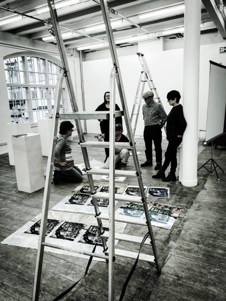 An A-frame ladder beside prints laid out on the gallery floor, with visitors in conversation, at Studio 44, Stockholm